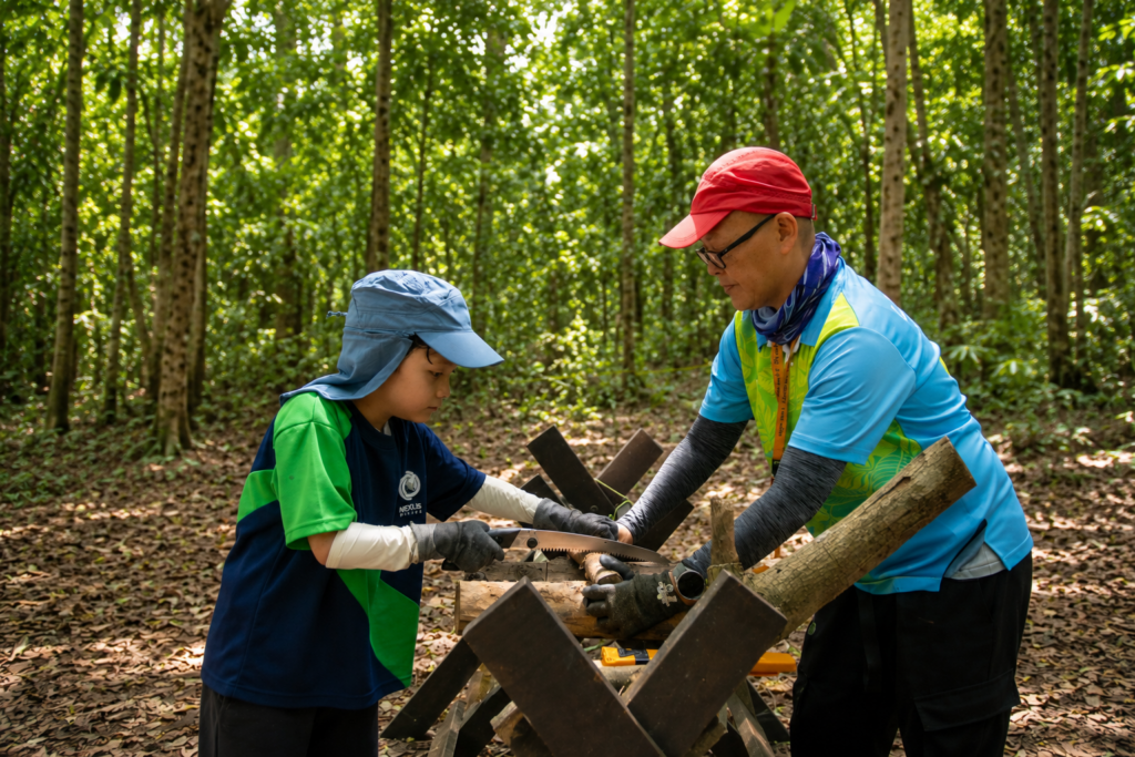 A Primary learner uses a hand saw with guidance from an FSLI-trained teacher during a Nexus Forest School outdoor learning session in the rainforest at Nexus International School Malaysia.