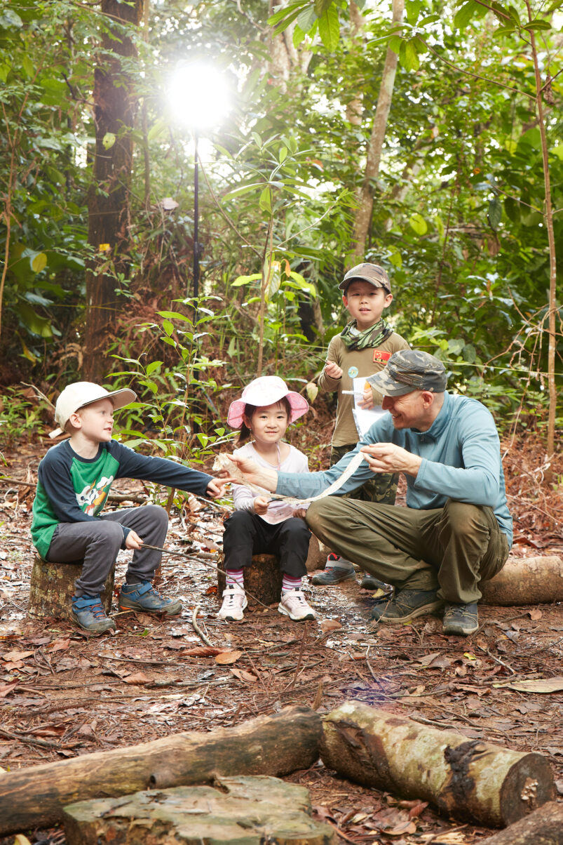 A Forest School educator and three young learners gathered in a forest, examining a natural object during an outdoor learning activity surrounded by lush greenery.
